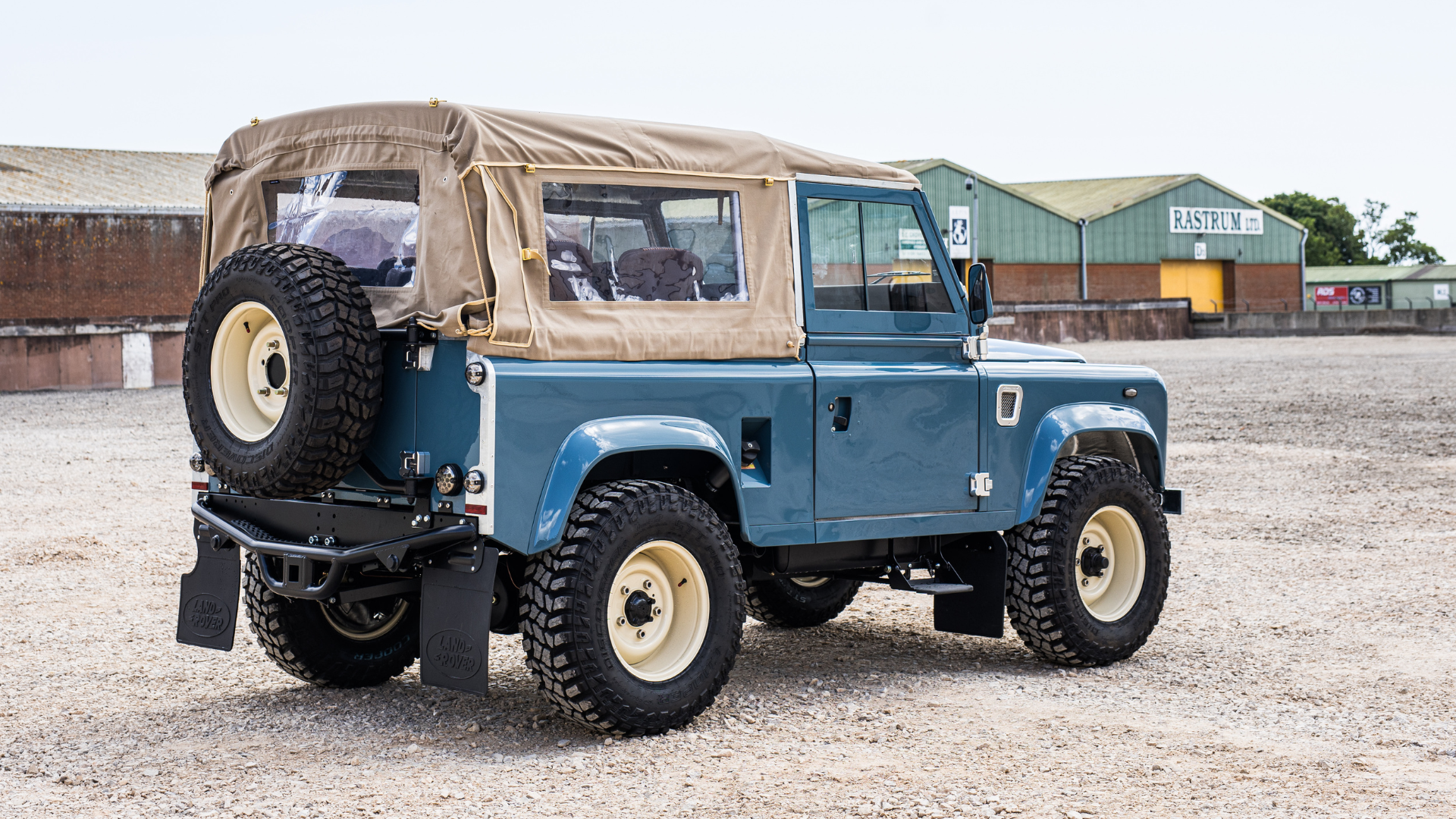 Land Rover Defender 90 soft top in blue with cream steel wheels, rear three-quarter view parked at a commercial yard