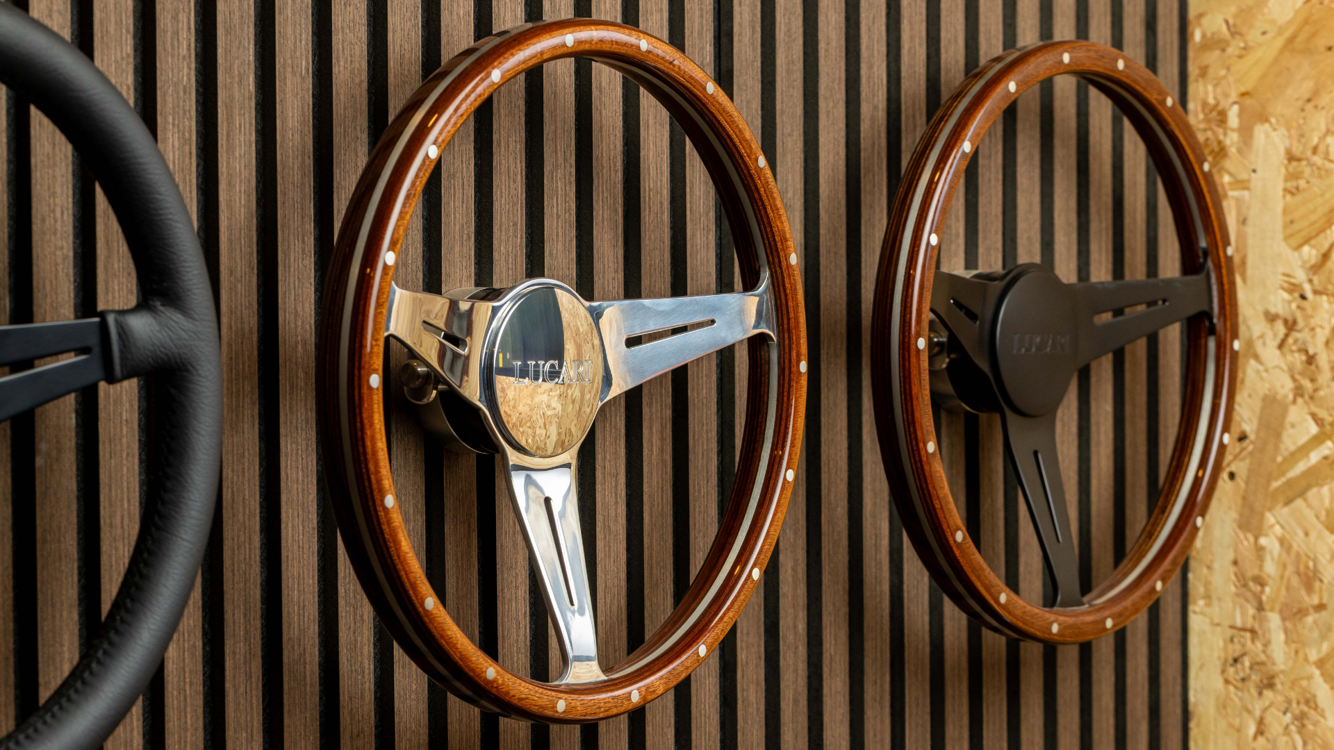 Three LUCARI steering wheels mounted on a slatted wood wall display: a black leather wheel on the left, a wooden-rim wheel with polished chrome three-spoke hub and LUCARI centre badge in the middle, and a matching wood-rim wheel with a black spoke hub on the right.