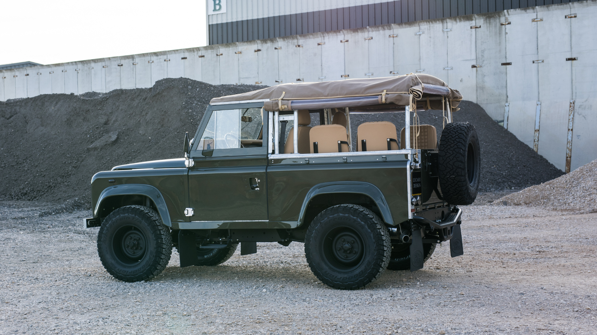 Side profile of a dark olive green Land Rover Defender 90 with a tan canvas soft top rolled back at the sides and rear, LUCARI Barley leather rear-inward seats visible and a rear-mounted spare wheel, parked on gravel with an industrial building behind.