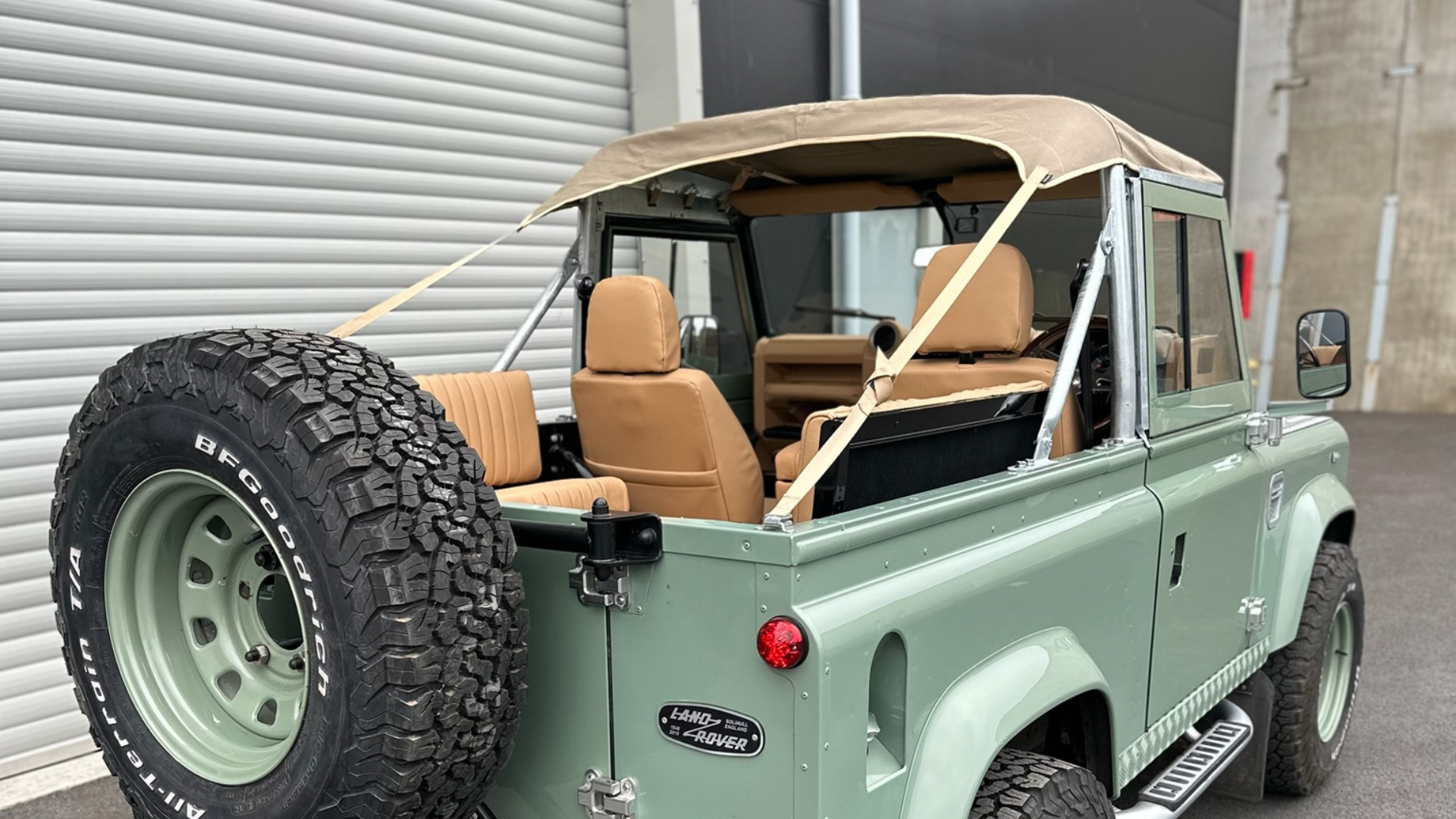 Rear three-quarter view of a sage green Land Rover Defender 90 with the Canvas Bikini Soft Top partially open and secured with tan straps, revealing LUCARI Barley leather interior seats. A BFGoodrich All-Terrain tyre on a matching sage green steel wheel is prominent in the foreground, photographed in front of roller shutter workshop doors.