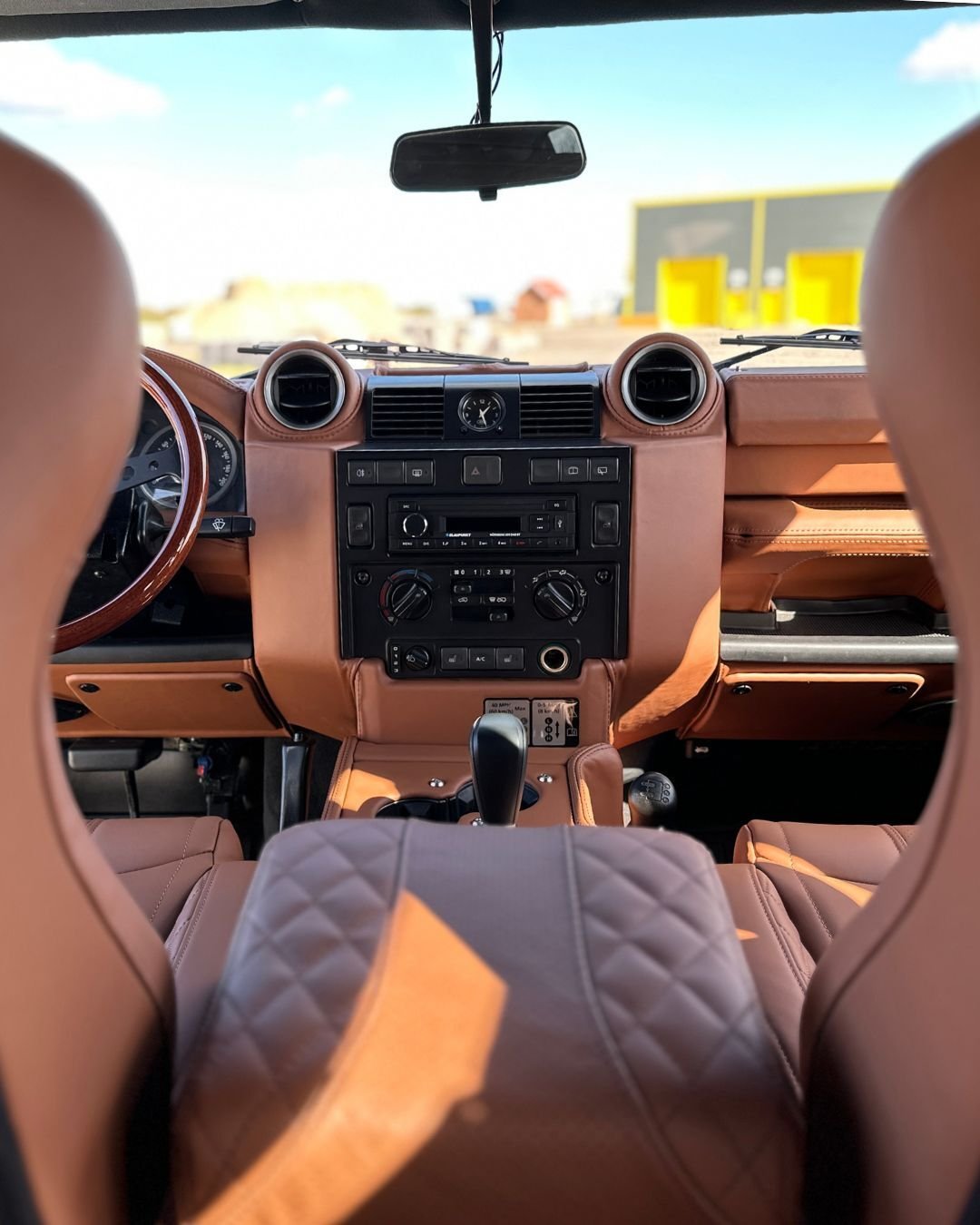 Interior view of a restored Land Rover Defender 110, featuring LUCARI Tan leather interior on the seats and door cards, a wooden steering wheel, and a custom auto cubby box. Sunshine through window highlights colour and texture throughout the cabin.