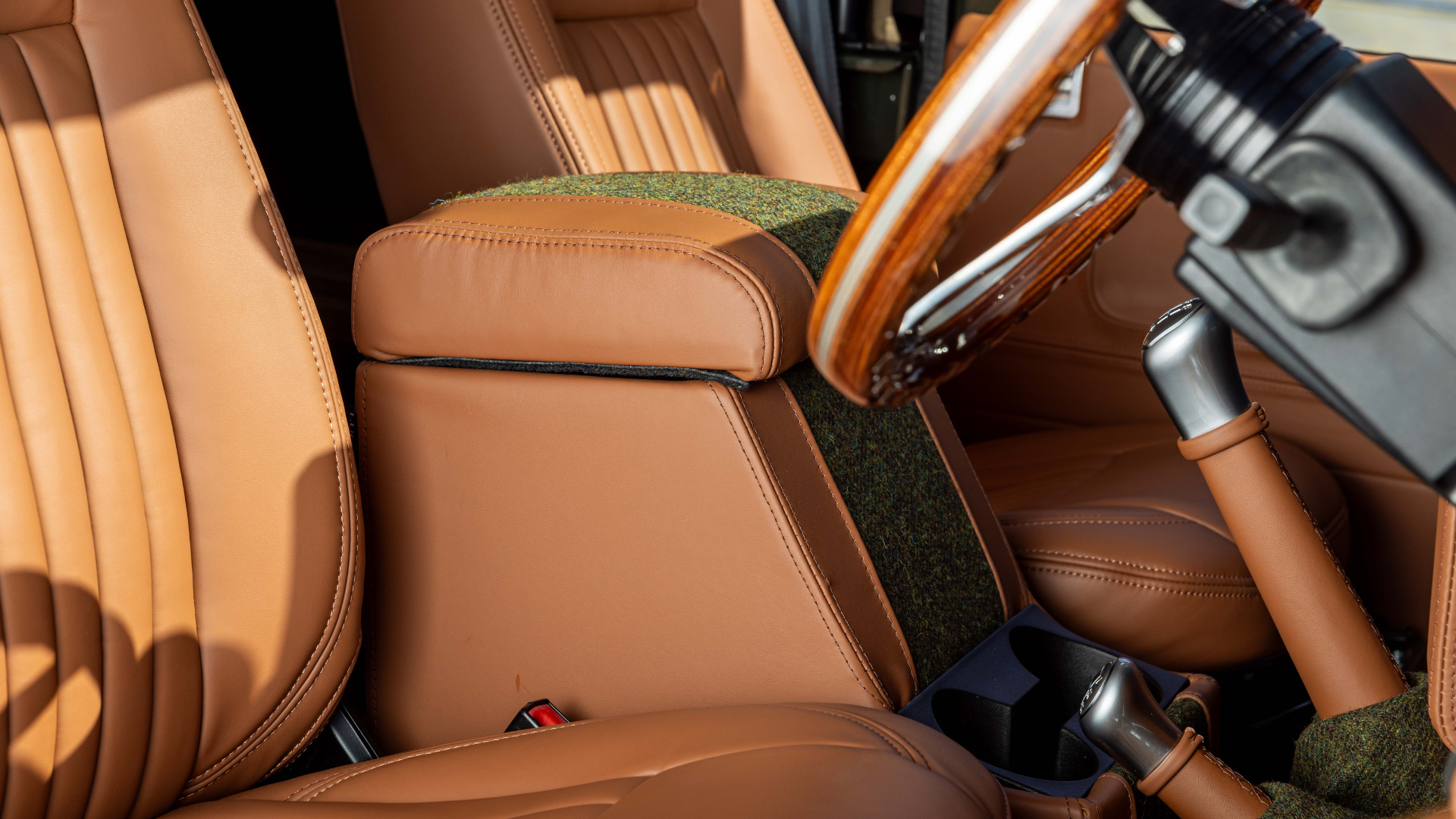Close-up of a Land Rover Defender interior showing a tan leather centre cubby box with Harris Tweed fabric inserts, a wood-rim steering wheel, and a chrome gear lever with a tan leather gaiter, photographed in warm natural light.