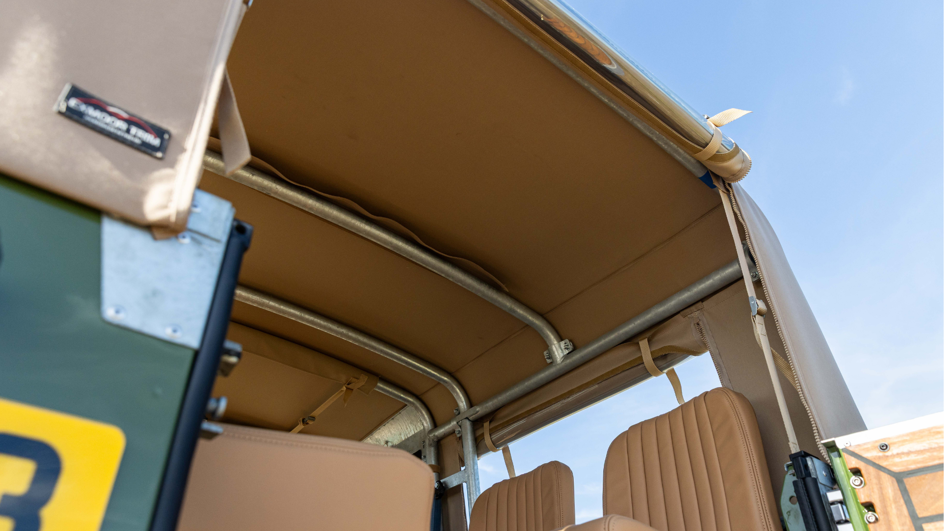 Upward view from inside a Land Rover Defender 90 showing the underside of a Sand Stayfast Soft Top stretched over galvanised roll cage hoops, with LUCARI Barley leather seats below and blue sky visible through the open sides.