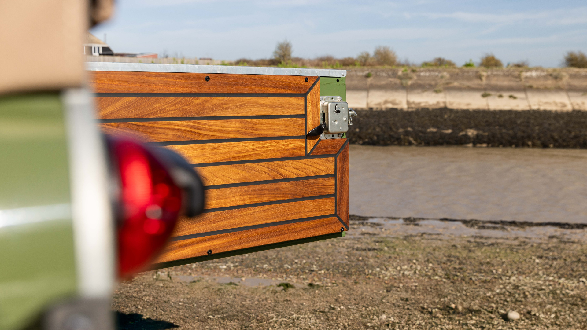 Close-up of an Iroko wood rear tailgate on a Land Rover Defender 90, showing the horizontal slat and black caulking detailing with a chrome latch fitting.