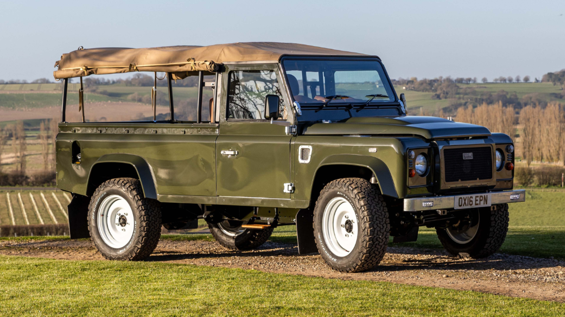 Green Defender 110 pickup with canvas tilt canopy and white steel wheels, parked on a hillside overlooking countryside.