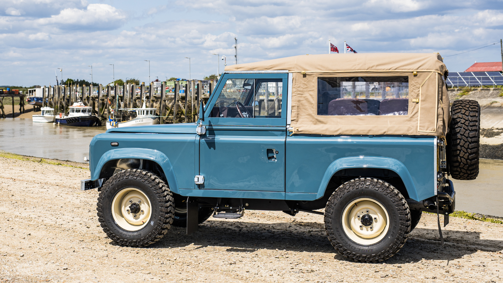 Side profile of a restored blue Land Rover Defender 90 fitted with a Sand Stayfast Soft Top with clear plastic side windows and cream steel wheels, and off-road tyres, parked on a shingle foreshore with a tidal harbour and moored boats in the background.