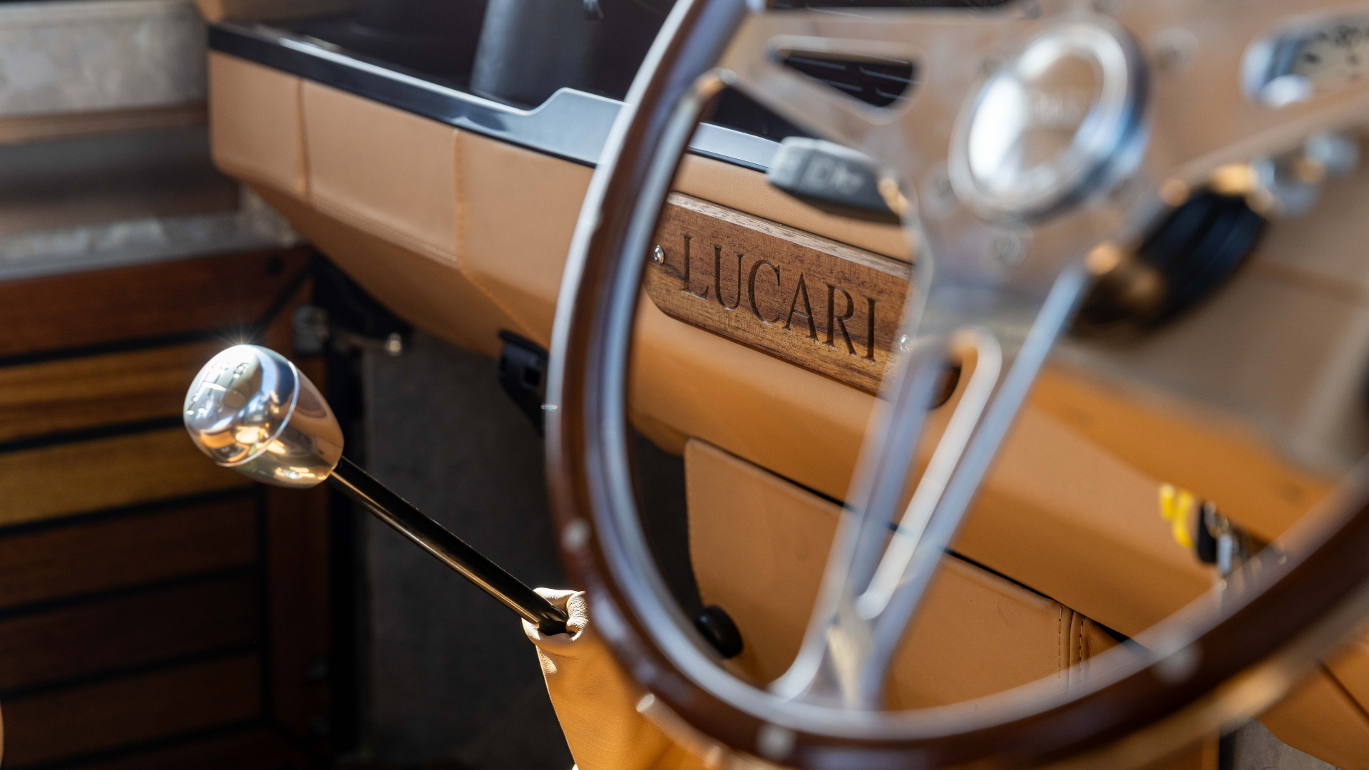 Interior detail of a Land Rover Defender showing a walnut wood fascia strip with the LUCARI name engraved into it, set against a tan leather dashboard, with a wood-rim steering wheel and chrome gear knob visible in the foreground.