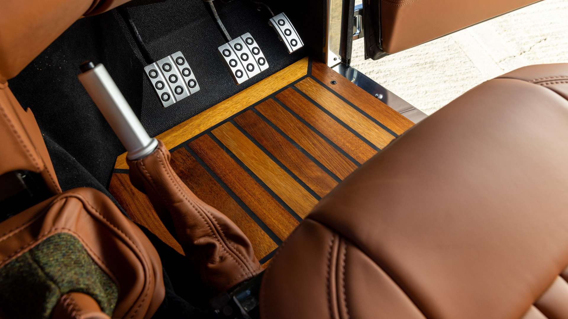 Overhead view of a Land Rover Defender footwell showing an Iroko wood floor panel, three polished aluminium drilled pedals on a black carpet surround, a tan leather gear gaiter, and Harris Tweed trim visible to the left alongside Tan leather seats.