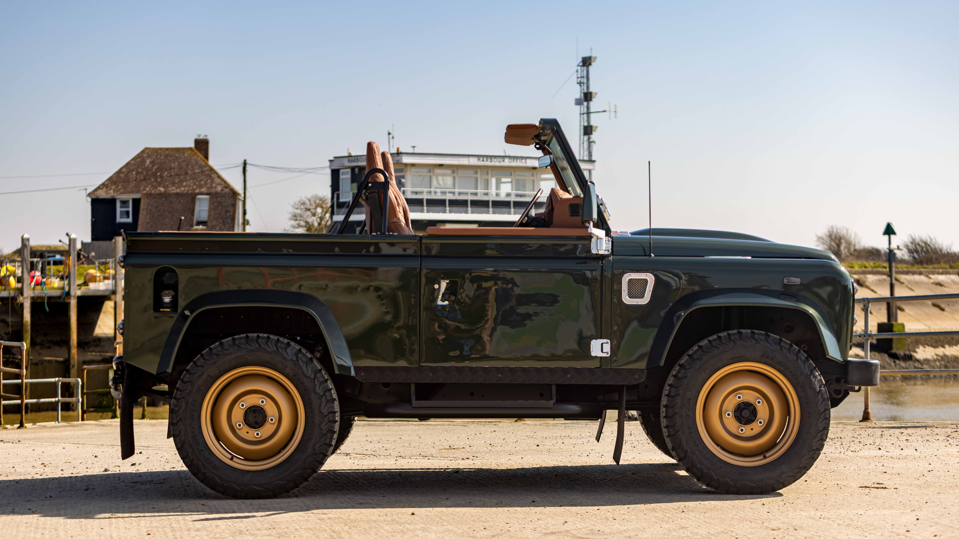 Side profile of a dark green Land Rover Defender 90 with the soft top fully removed, showing tan leather seats and half doors, fitted with gold steel wheels and off-road tyres, parked on a harbourside quay with the harbour office building visible behind.