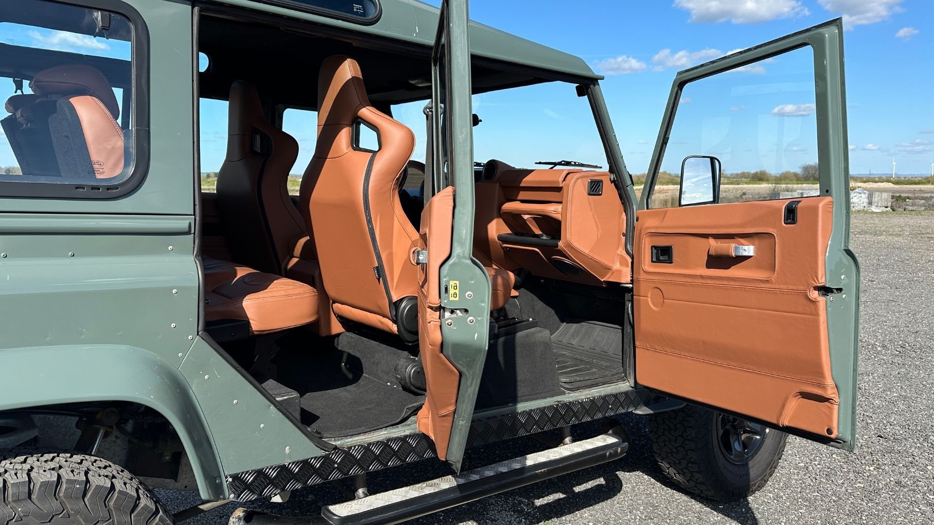Rear side doors of a green Land Rover Defender 110 open wide, revealing a full LUCARI Tan leather interior with diamond-stitched bucket seats, matching door cards, and chequer plate floor detailing. Blue sky and open landscape visible in the background.