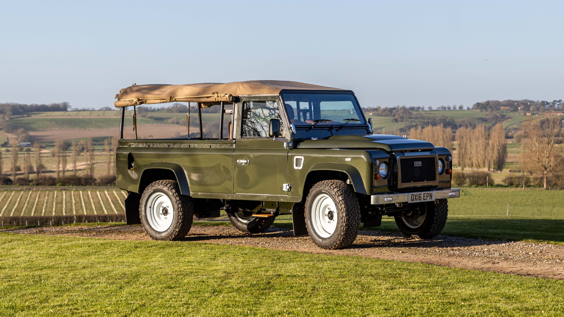 An olive green Land Rover Defender 110 with a sand-coloured canvas hood rolled back, white steel wheels, and off-road tyres, parked on a gravel track surrounded by manicured lawn and open English countryside