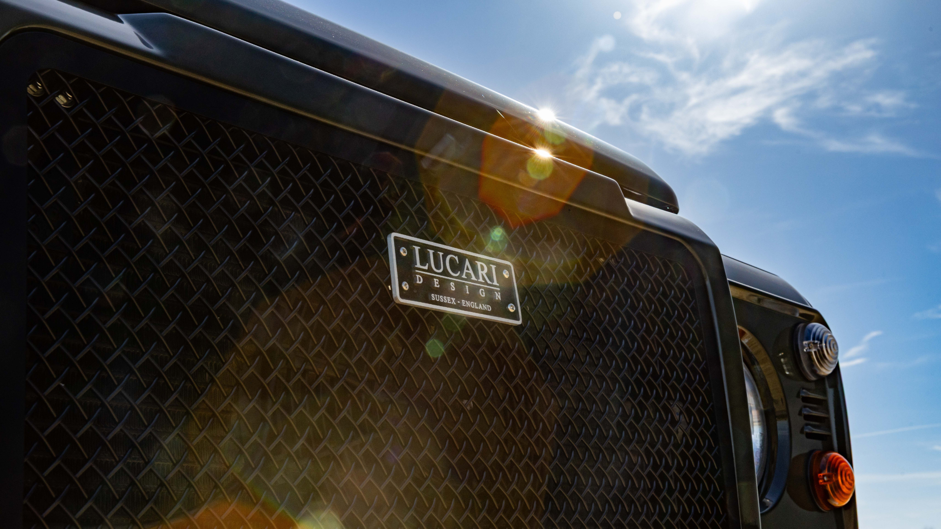 Close-up of a LUCARI Design, Sussex, England badge mounted on the black diamond-plate front grille of a Land Rover Defender, shot against a bright blue sky with sun flare.