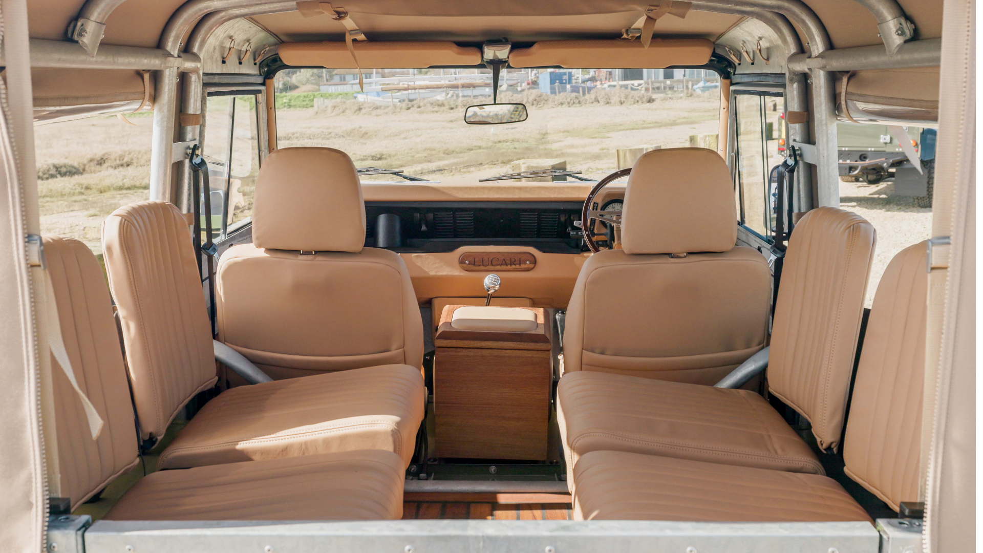 Interior view of a Land Rover Defender shot from the rear loadspace, showing four sand leather rear inward seats with fluted stitch detailing, a wooden centre console with a chrome gear knob, a LUCARI-branded dashboard fascia, and a LUCARI Iroko rear deck floor, with the canvas hood frame visible overhead.