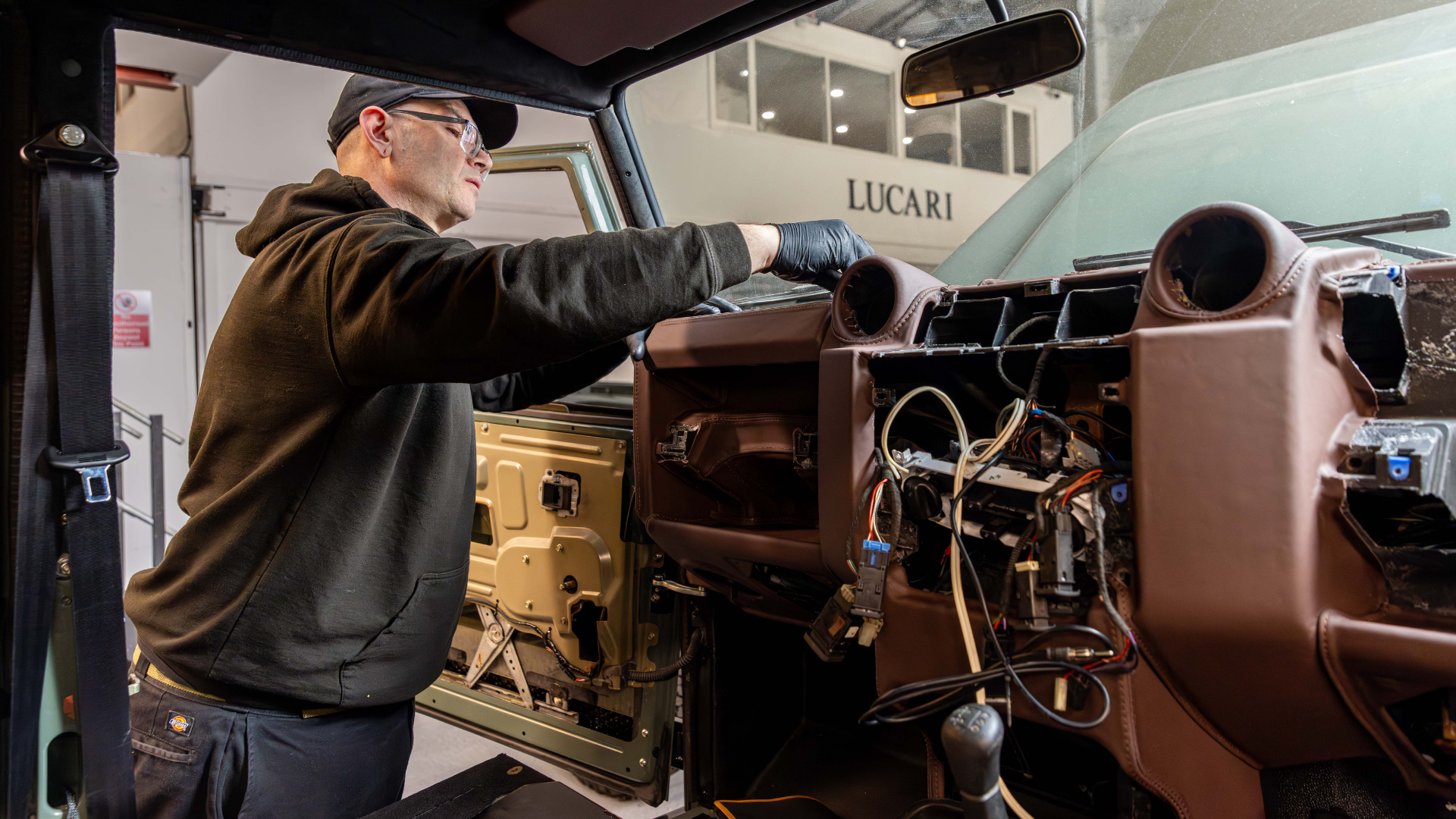 A LUCARI Install Technician wearing black gloves works on the dashboard and wiring of a Land Rover Defender mid-build, with the LUCARI workshop signage visible through the windscreen in the background.
