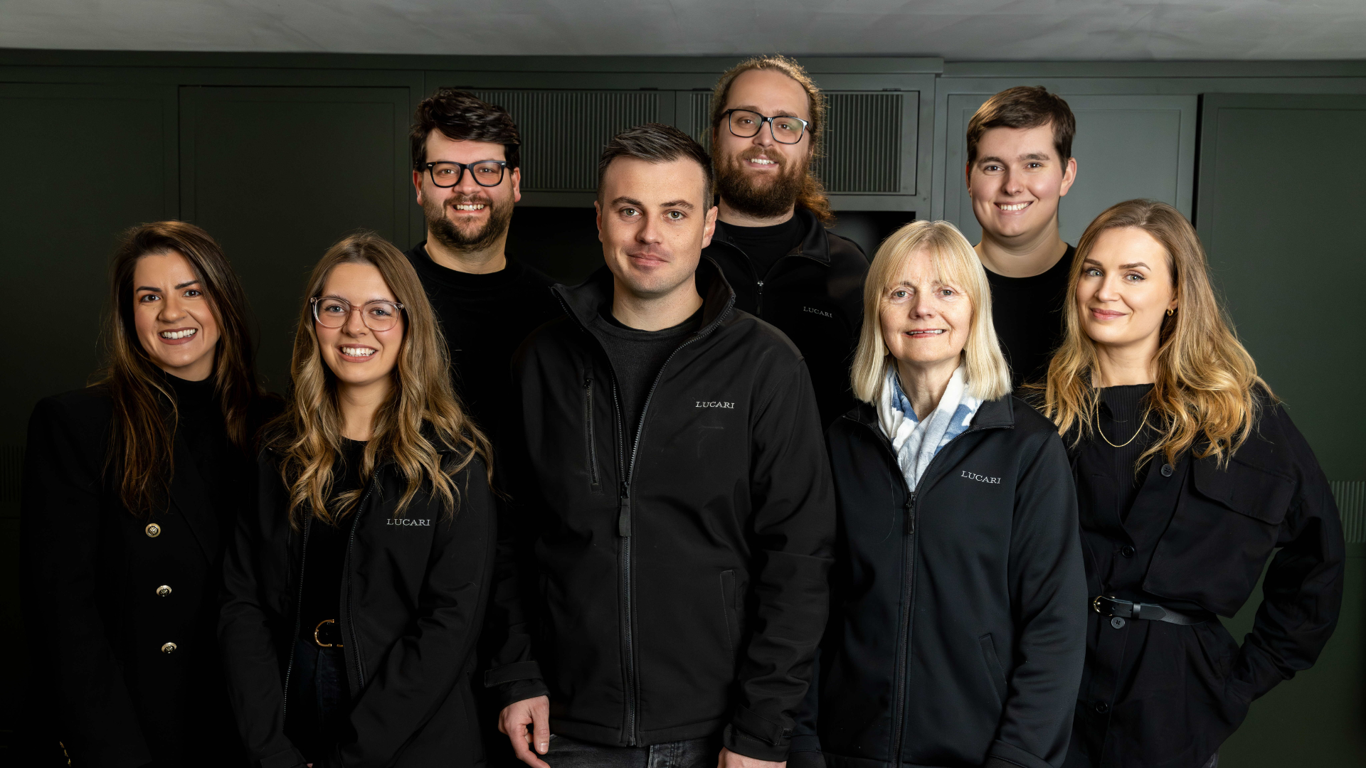 The LUCARI Retail team of eight posed together in front of a dark olive green panelled wall