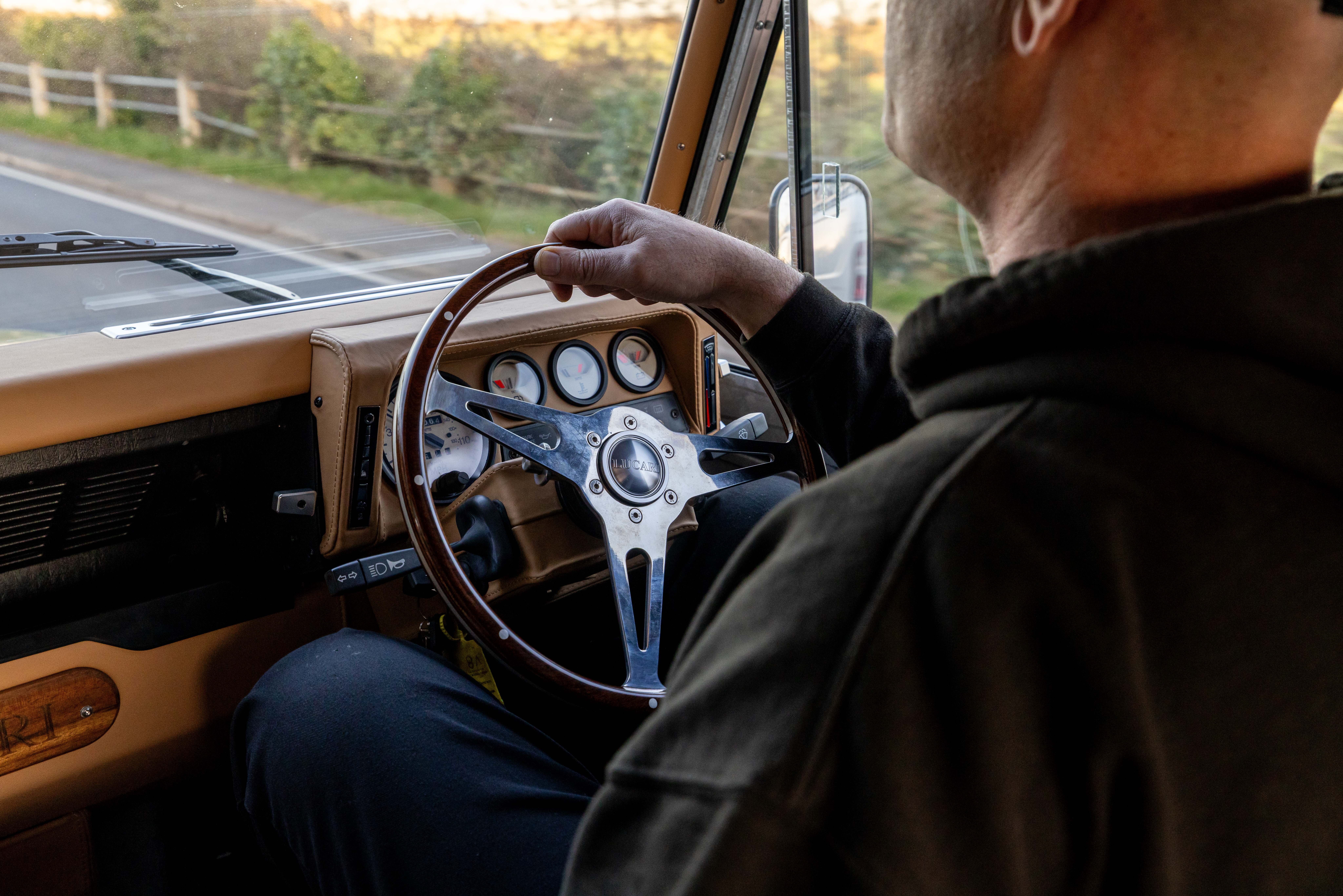 Driver's over-the-shoulder view inside a LUCARI-built Land Rover Defender, showing a LUCARI wooden dished steering wheel with LUCARI centre badge and barley leather dashboard trim