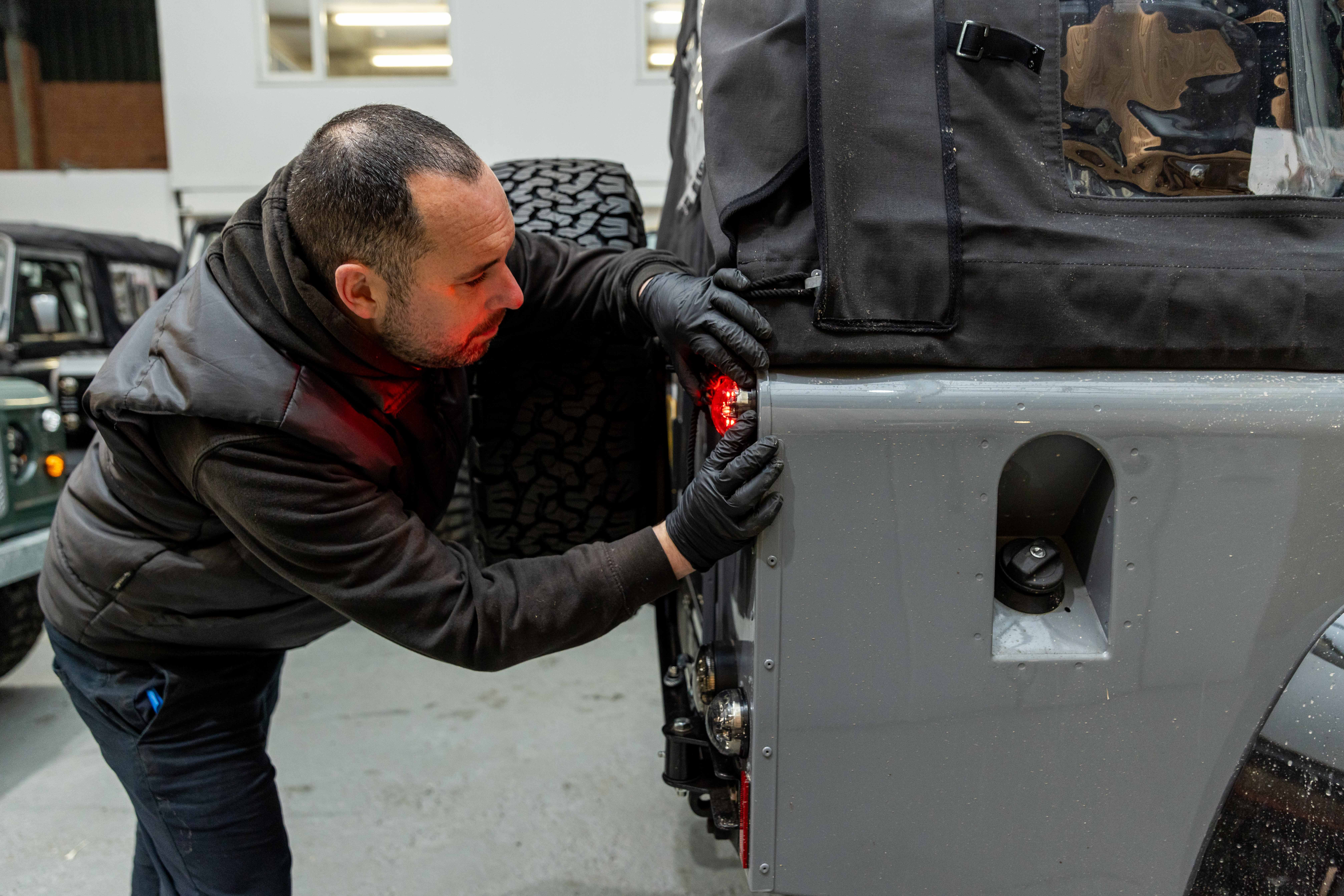A LUCARI technician in black gloves leaning in closely to fit a rear light unit to a silver Land Rover Defender, with a black canvas soft top and spare wheel visible above and another Defender partially visible in the background.