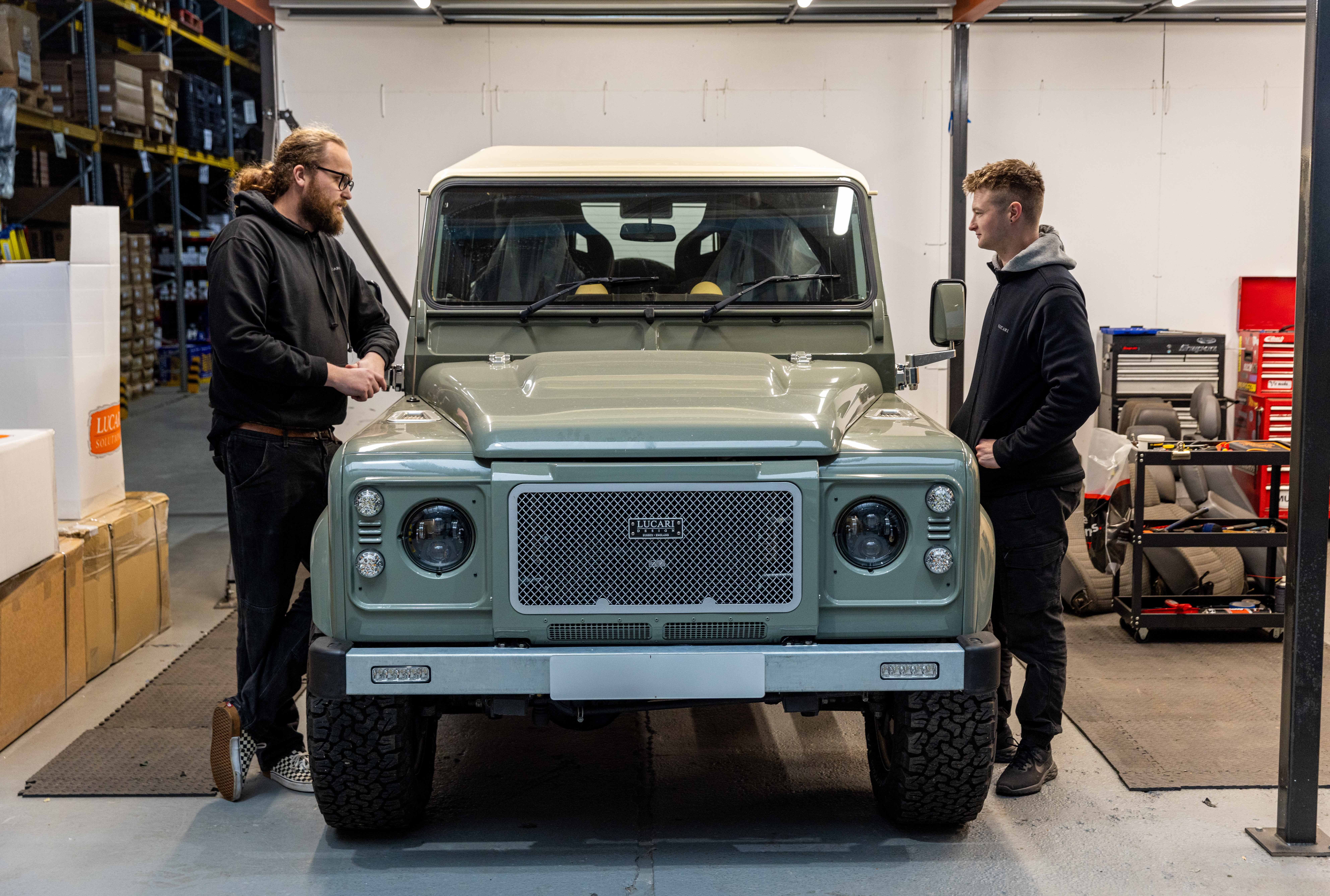 Two LUCARI team members standing either side of a sage green Land Rover Defender 90 fitted with a Lucari Design mesh grille and LED headlights, discussing the build inside the LUCARI warehouse facility.