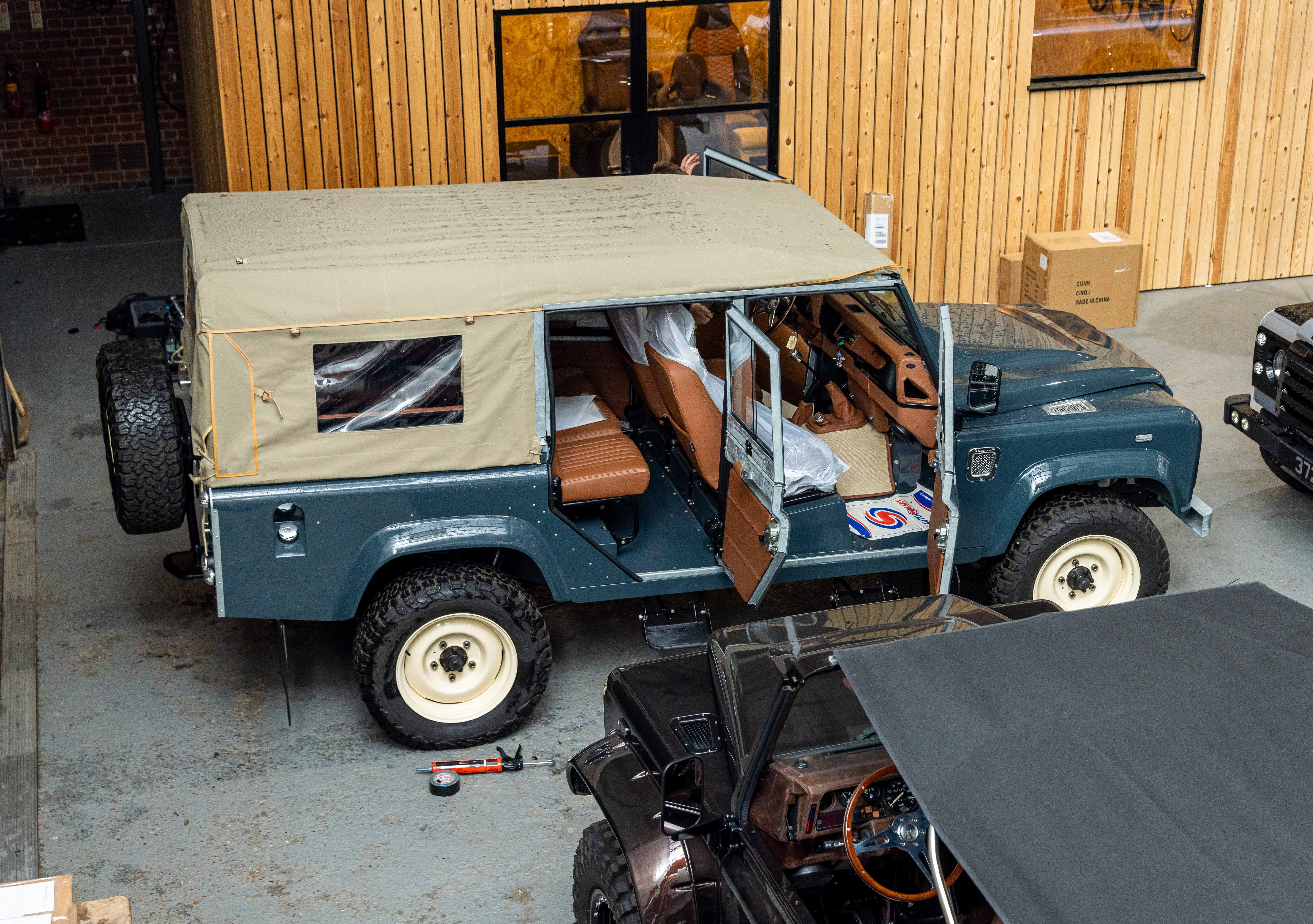 Elevated view looking down into the LUCARI workshop showing a teal blue Land Rover Defender 110 with all doors open, fitted with a sand canvas soft top and cream steel wheels, with tan leather interior visible and another dark Defender partially in frame at the bottom.