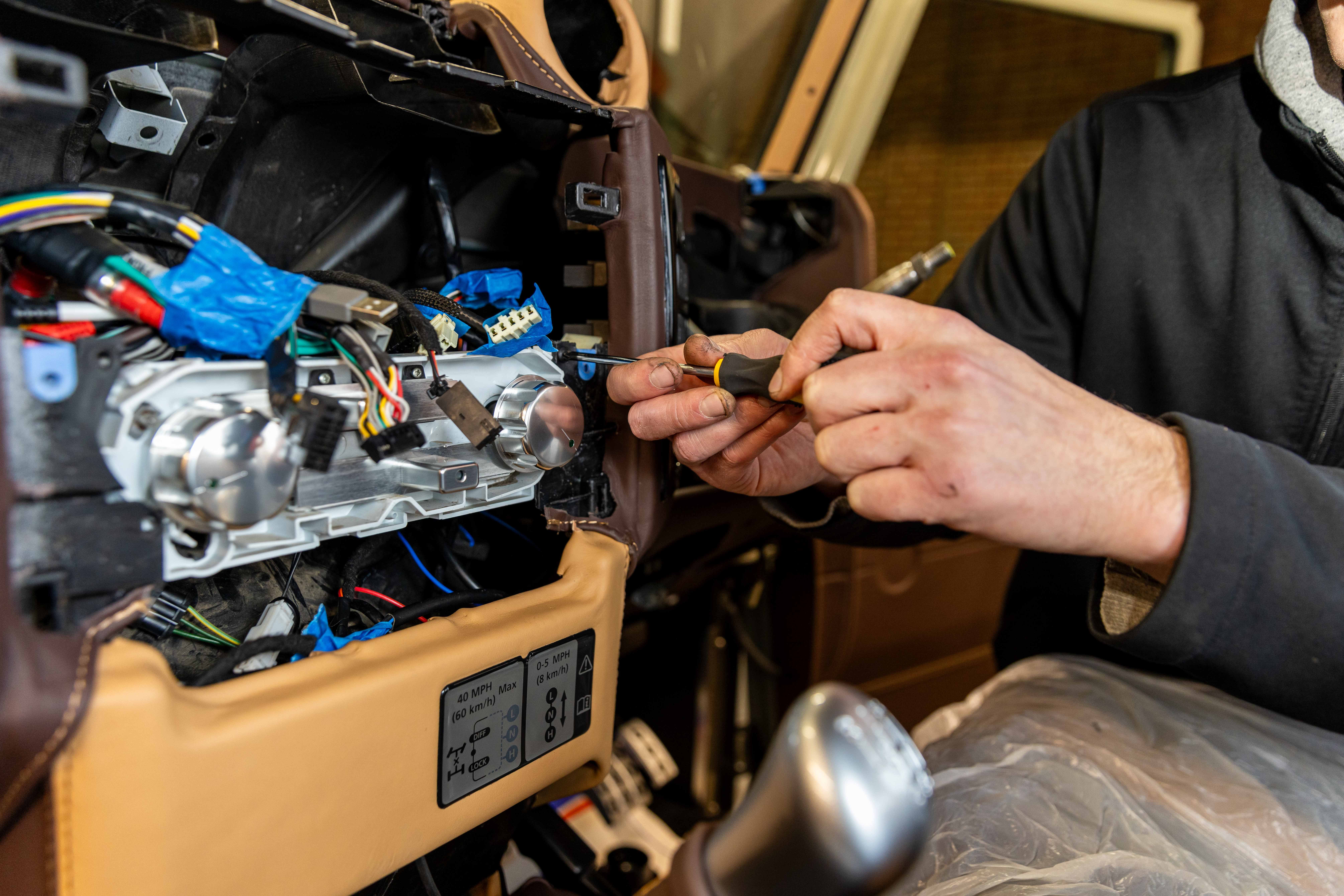 Close-up of a LUCARI technician using a screwdriver to work on the wiring loom and instrument cluster behind a leather-wrapped Land Rover Defender dashboard, with coloured wiring, connectors, and chrome switch components visible in detail.