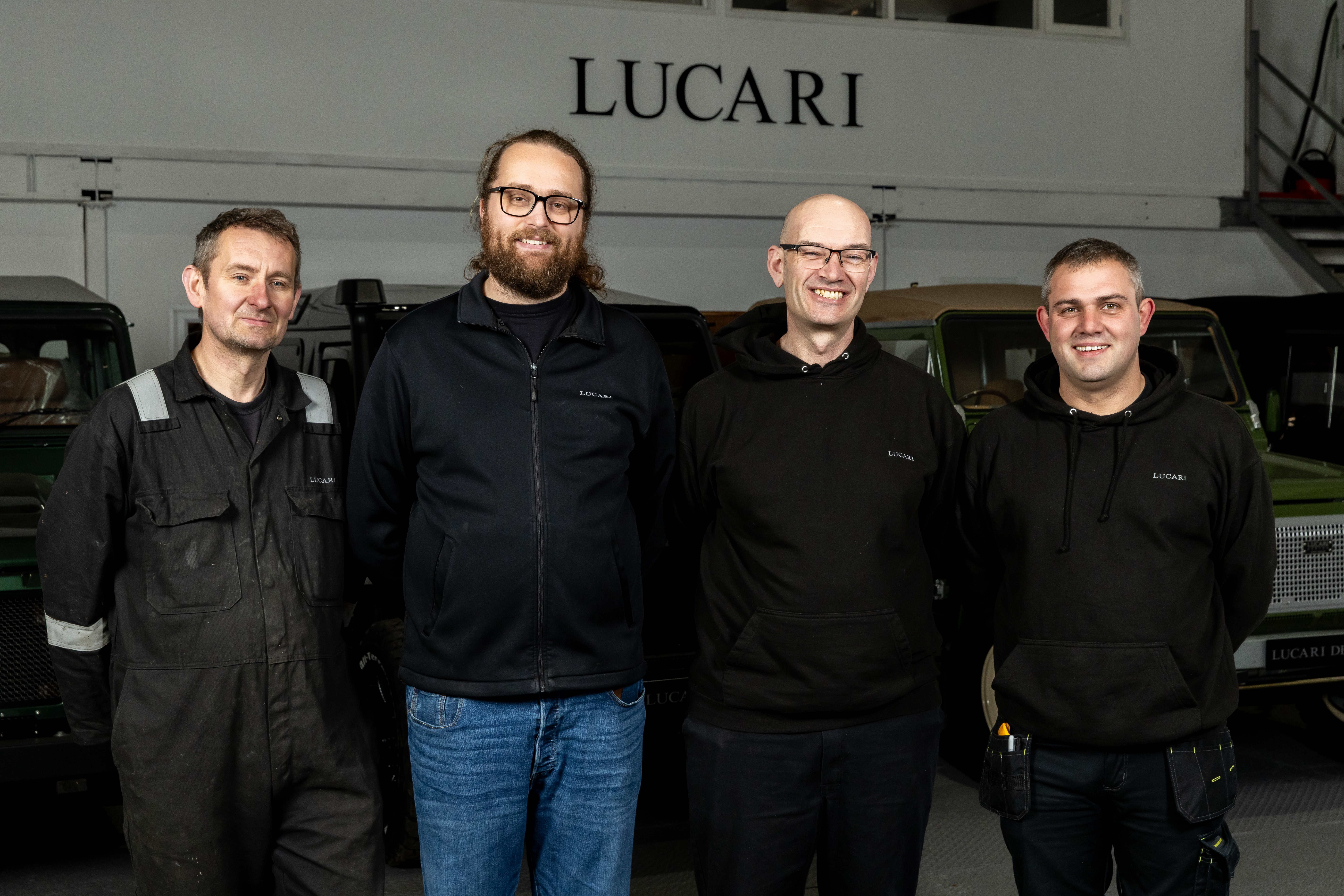 Four members of the LUCARI fitting and installation team standing together in front of the LUCARI workshop, smiling at the camera. All four are wearing branded LUCARI workwear, with Land Rover Defenders and the LUCARI workshop signage visible behind them.