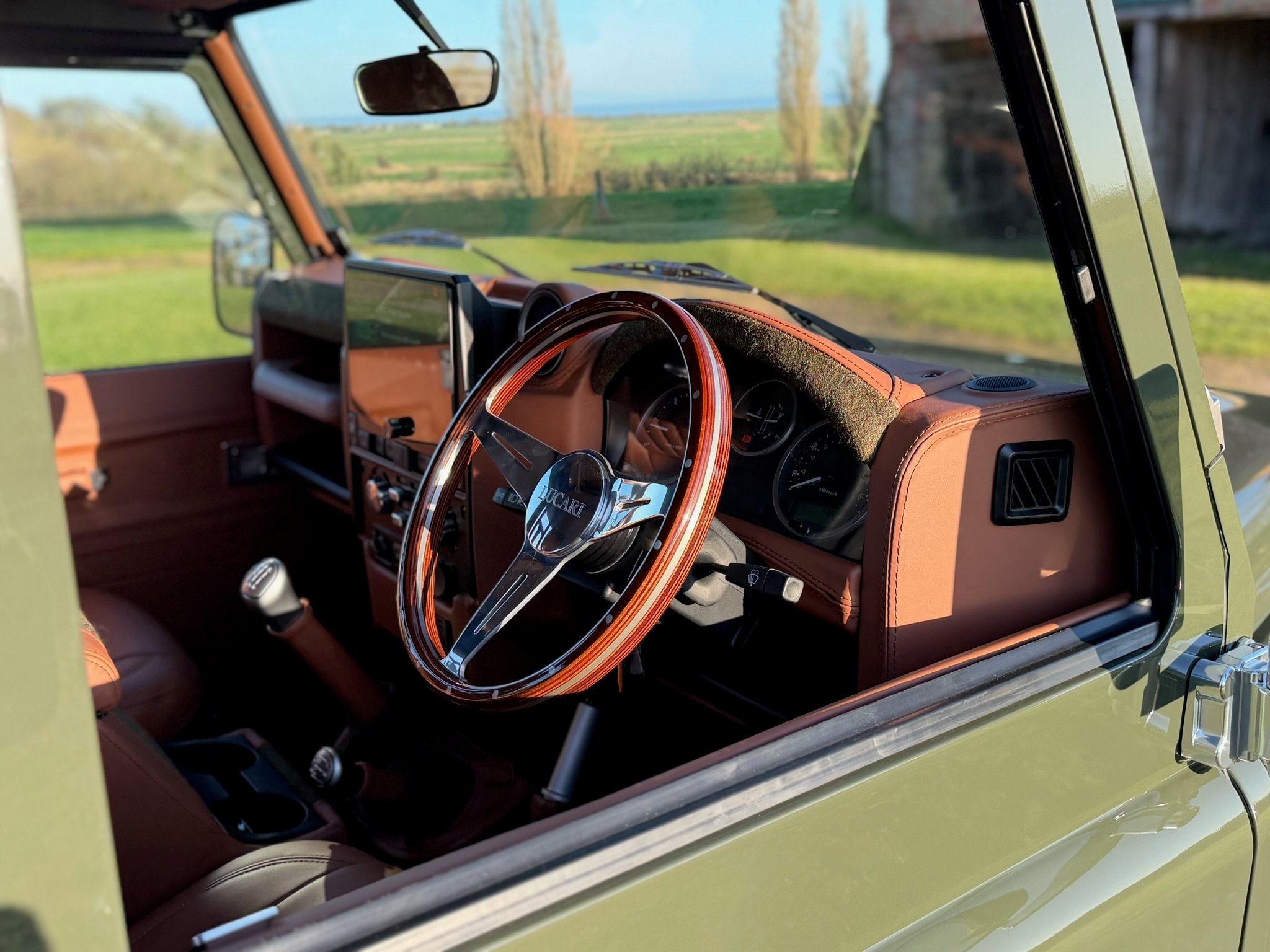 Interior view through the open window of a LUCARI-built Land Rover Defender 110, showing a LUCARI wooden dished steering wheel, Tan leather trim throughout and a countryside backdrop