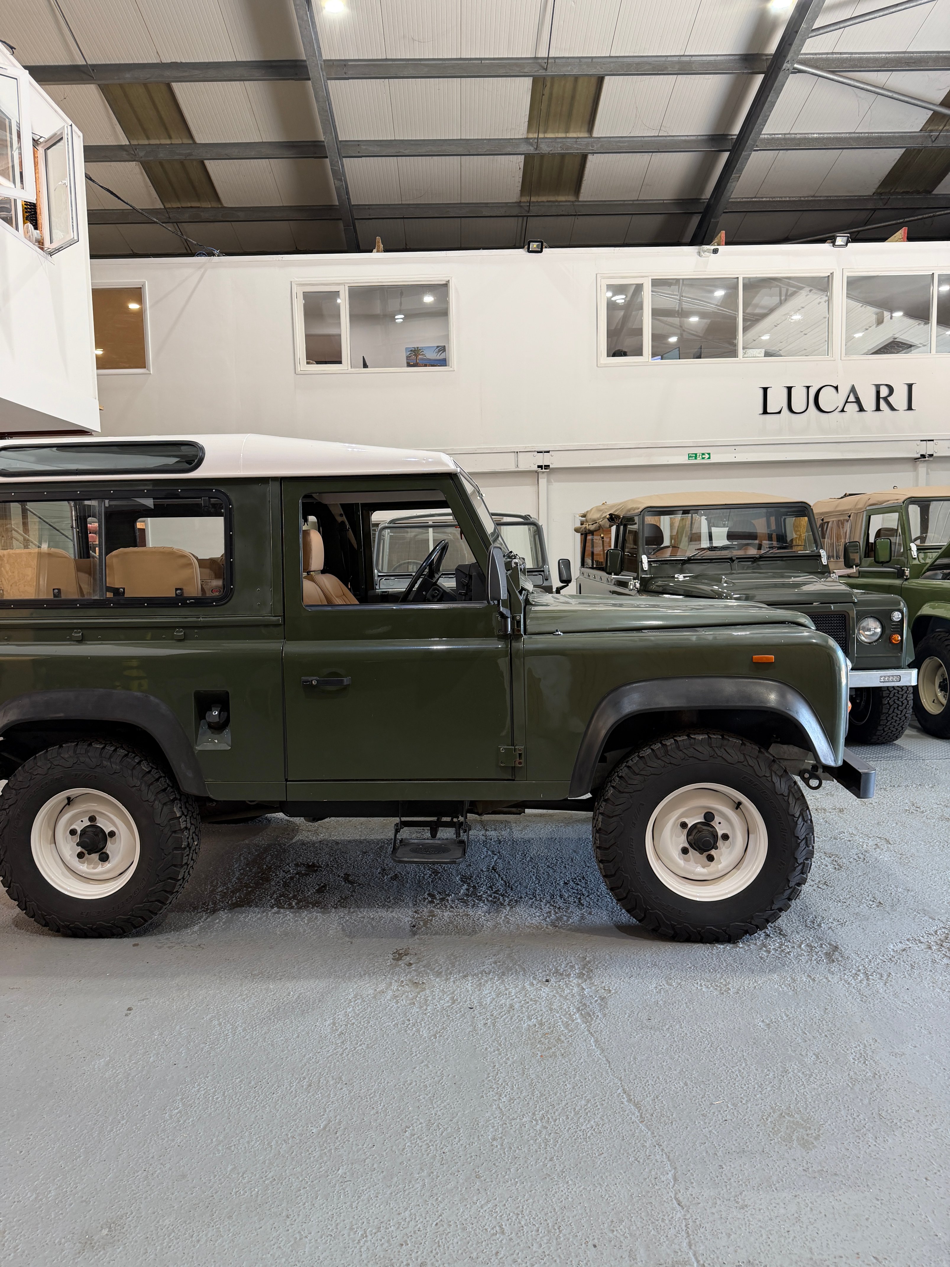 Side profile of an olive green Land Rover Defender 90 with a white hard top and cream steel wheels, parked inside the LUCARI workshop with additional Defenders and the LUCARI workshop signage visible in the background.