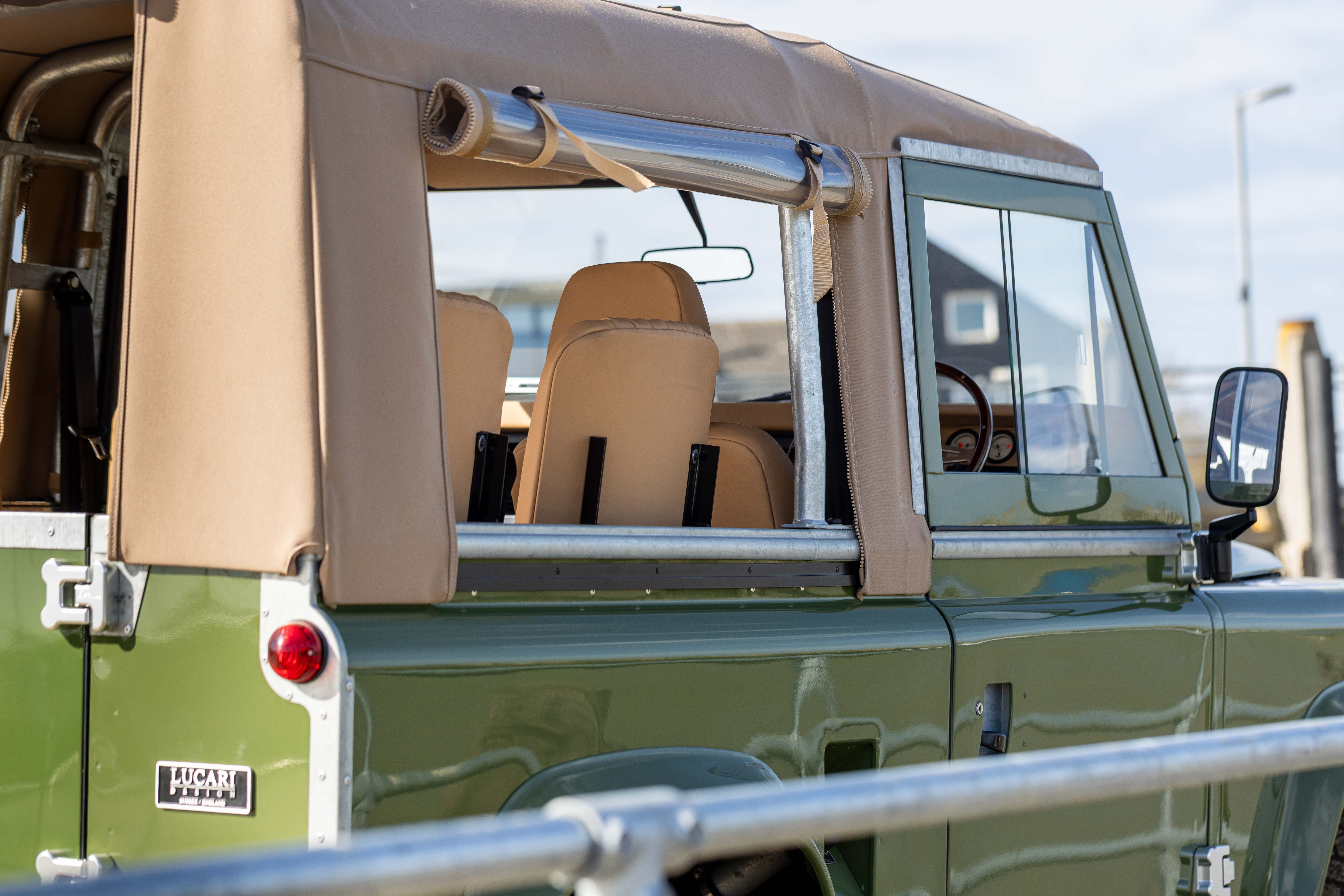 Close-up of a Sand Stayfast soft top on a Keswick Green Land Rover Defender 90, showing the rolled front section on a polished aluminium rail, clear side windows, and tan leather seats visible through the open rear section.