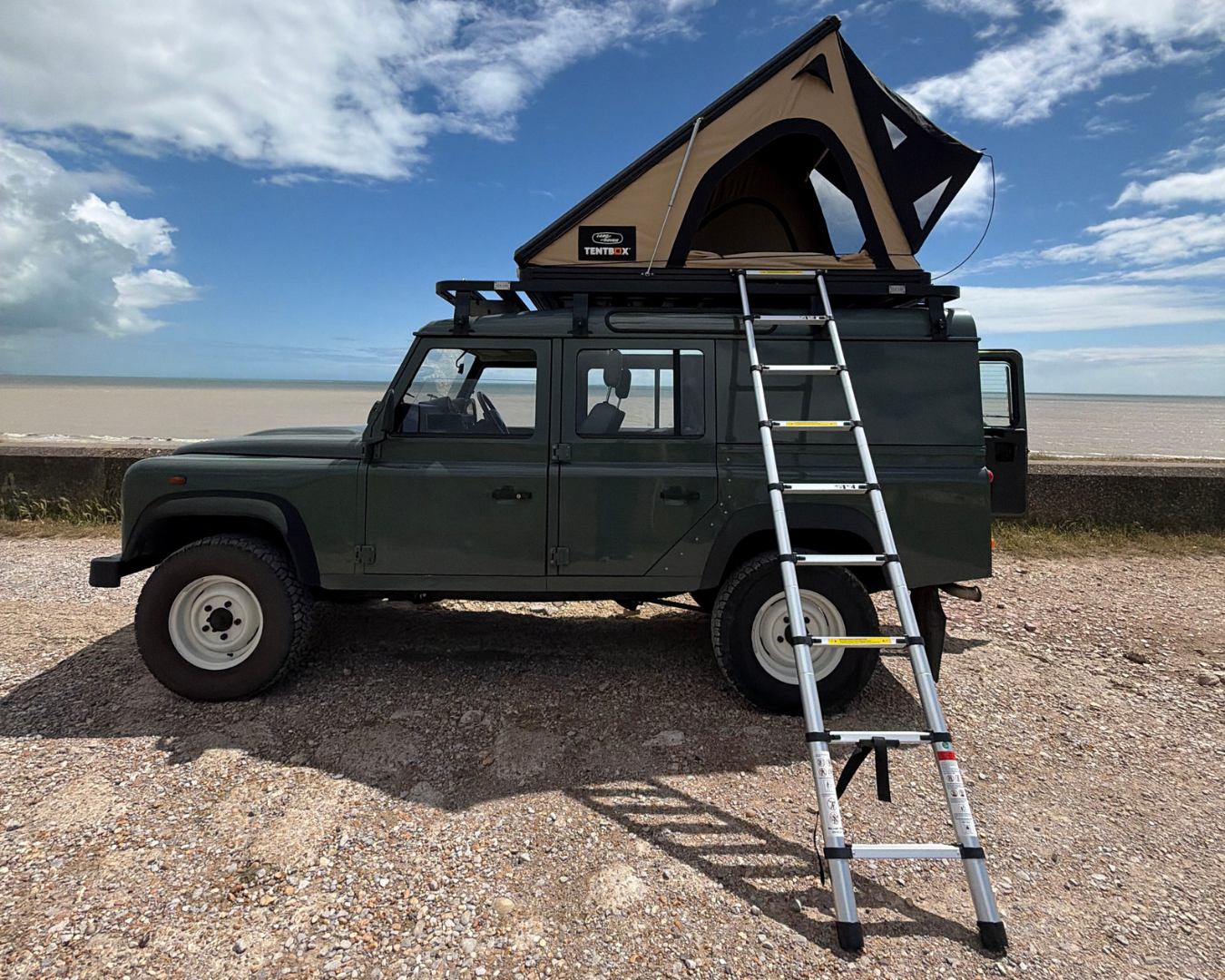 Land Rover Defender 110 with TentBox Cargo, Land Rover edition, open and ladder deployed, on top of a LUCARI Defender 110 Roof Rack, parked on coastal road