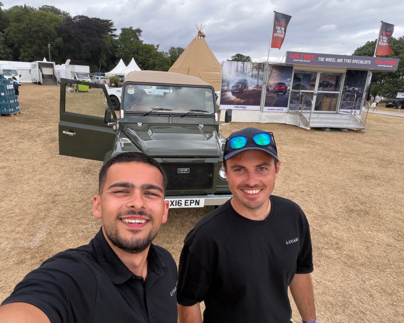 Two LUCARI team members taking a selfie in front of a LUCARI rebuilt Land Rover Defender 110 at The Game Fair, with a marquee tent and 4x4 Tyres stand in the background.