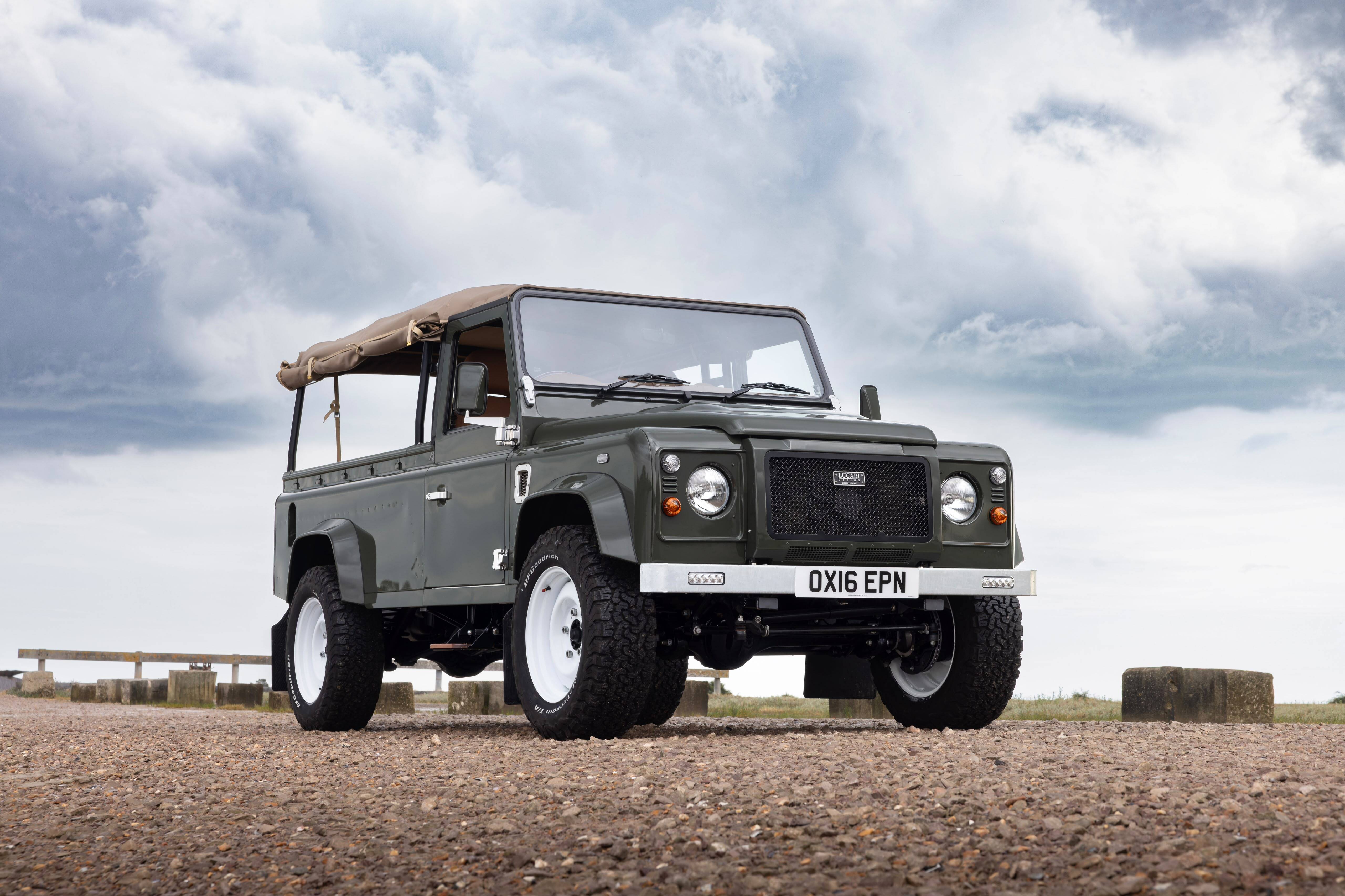 Land Rover Defender 110 pickup with canvas tilt canopy and LUCARI badged grille, front three-quarter view on a coastal gravel path.